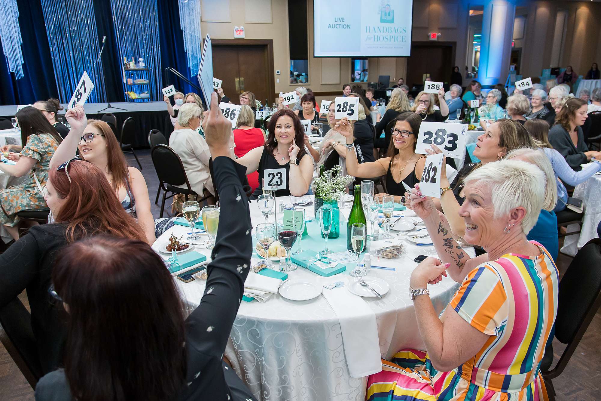 A large group of women seated in a banquet hall bidding on auction items at Handbags for Hospice