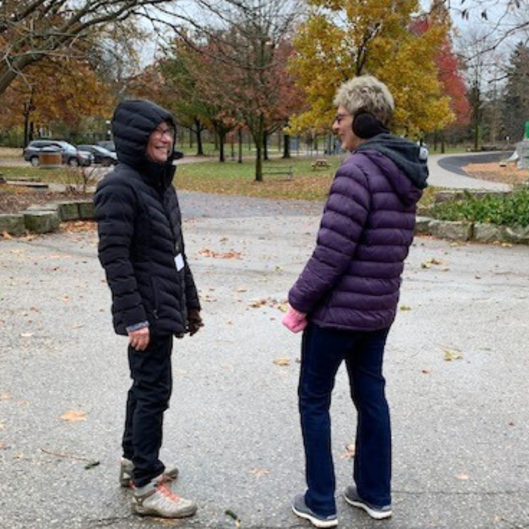 Two woman stand smiling at each other in a parking lot. They are dressed for cold weather.