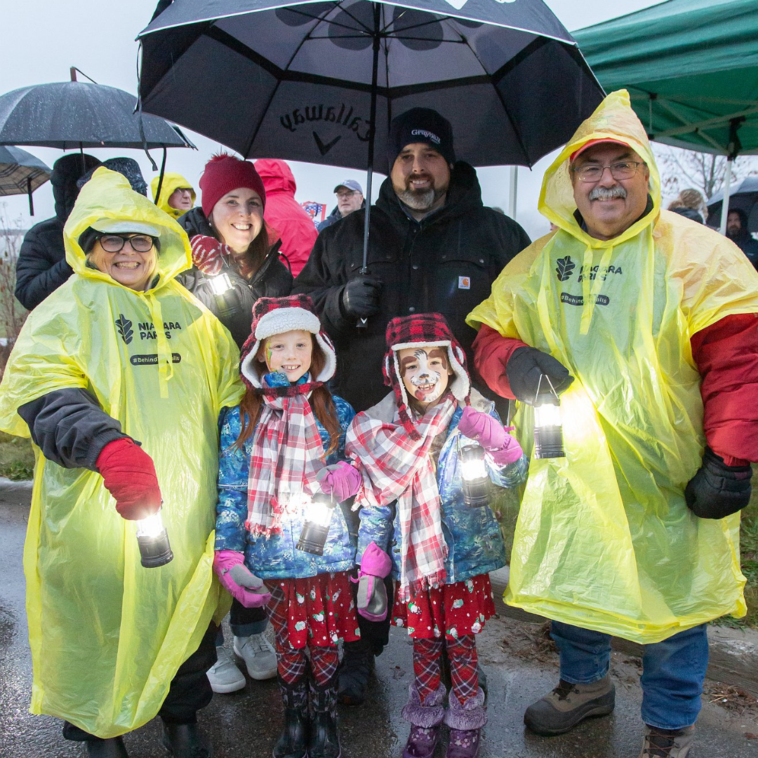 A group of people pose for a photo. It is raining and they are holding umbrellas and wearing rain gear.