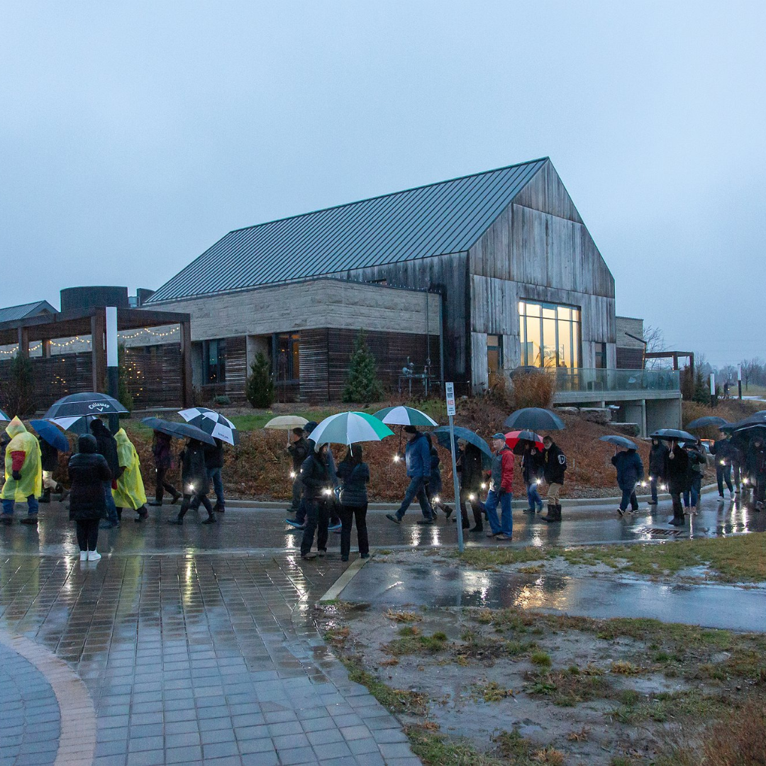 A group of people carrying lanterns and umbrellas walks along the back side of The Gies Family Centre.