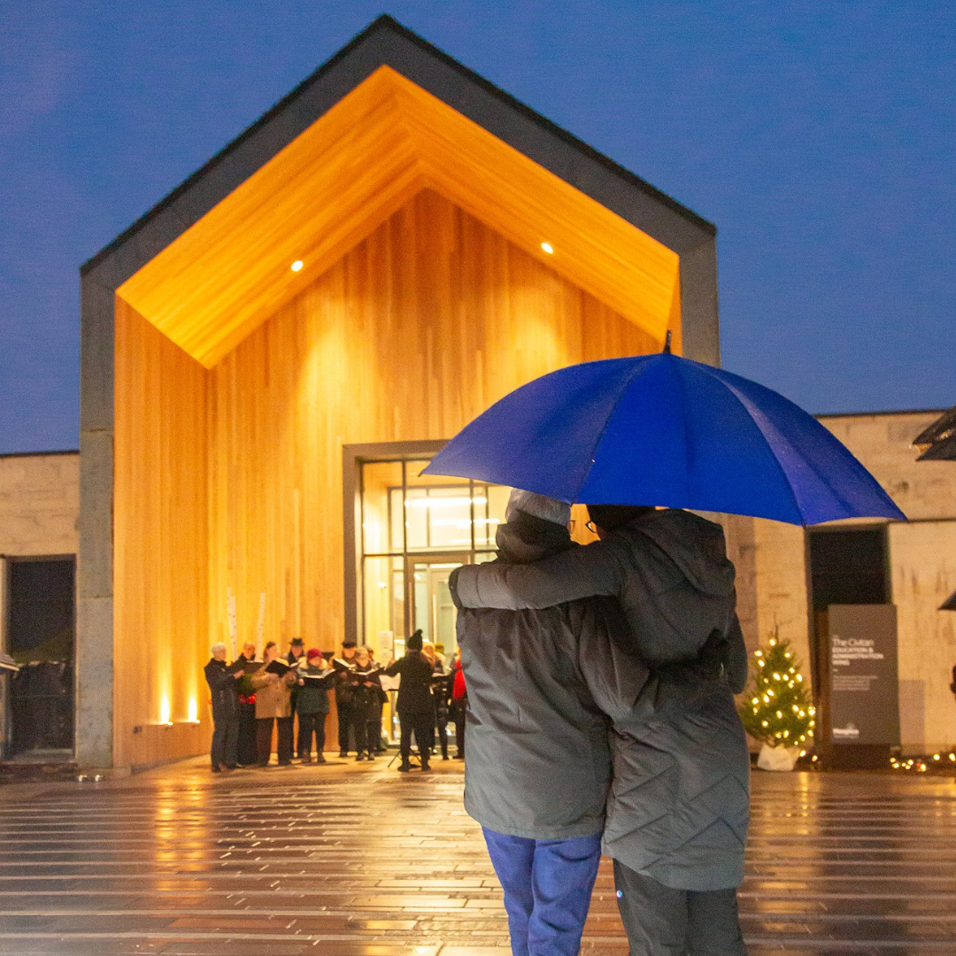 Two people stand together in an embrace. They are holding a blue umbrella and standing in front of The Gies Family Centre