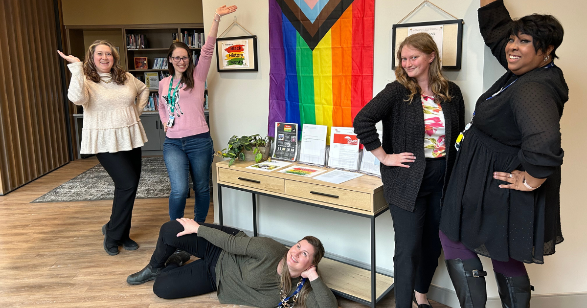 A group of five women pose for a photo. Four are standing, and some have an arm in the air. One is laying down on the floor with her hand on her hip. There is a table and an 2SLGBTQ+ flag in the middle of the photo.